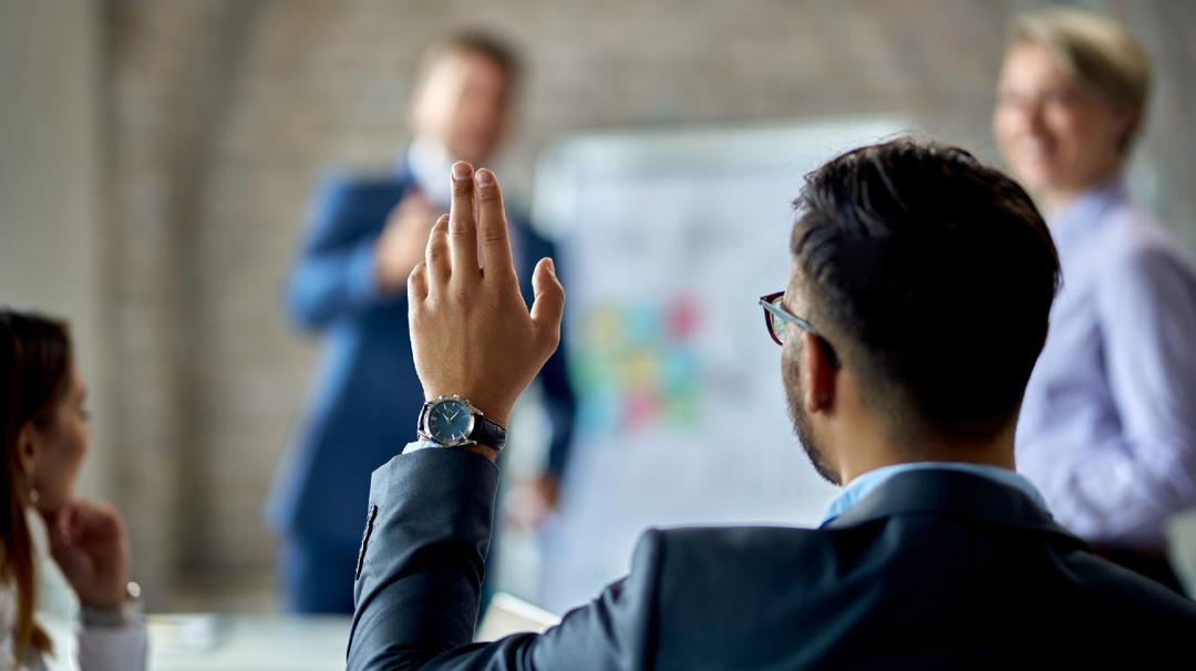 A man in a business suit raising his hand during a meeting with colleagues gathered around a whiteboard in a modern office.