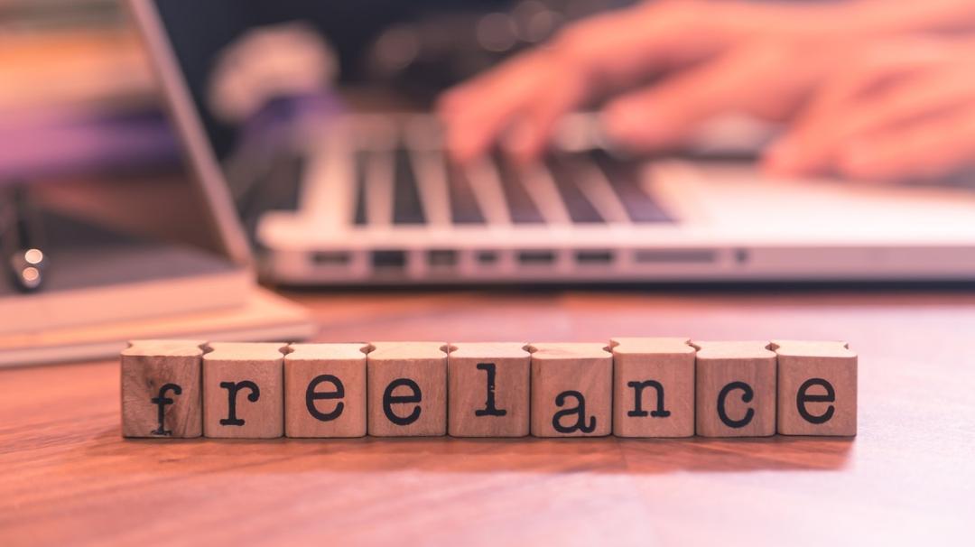 Wooden blocks spelling "freelance" on a desk with a blurred background featuring a person typing on a laptop.